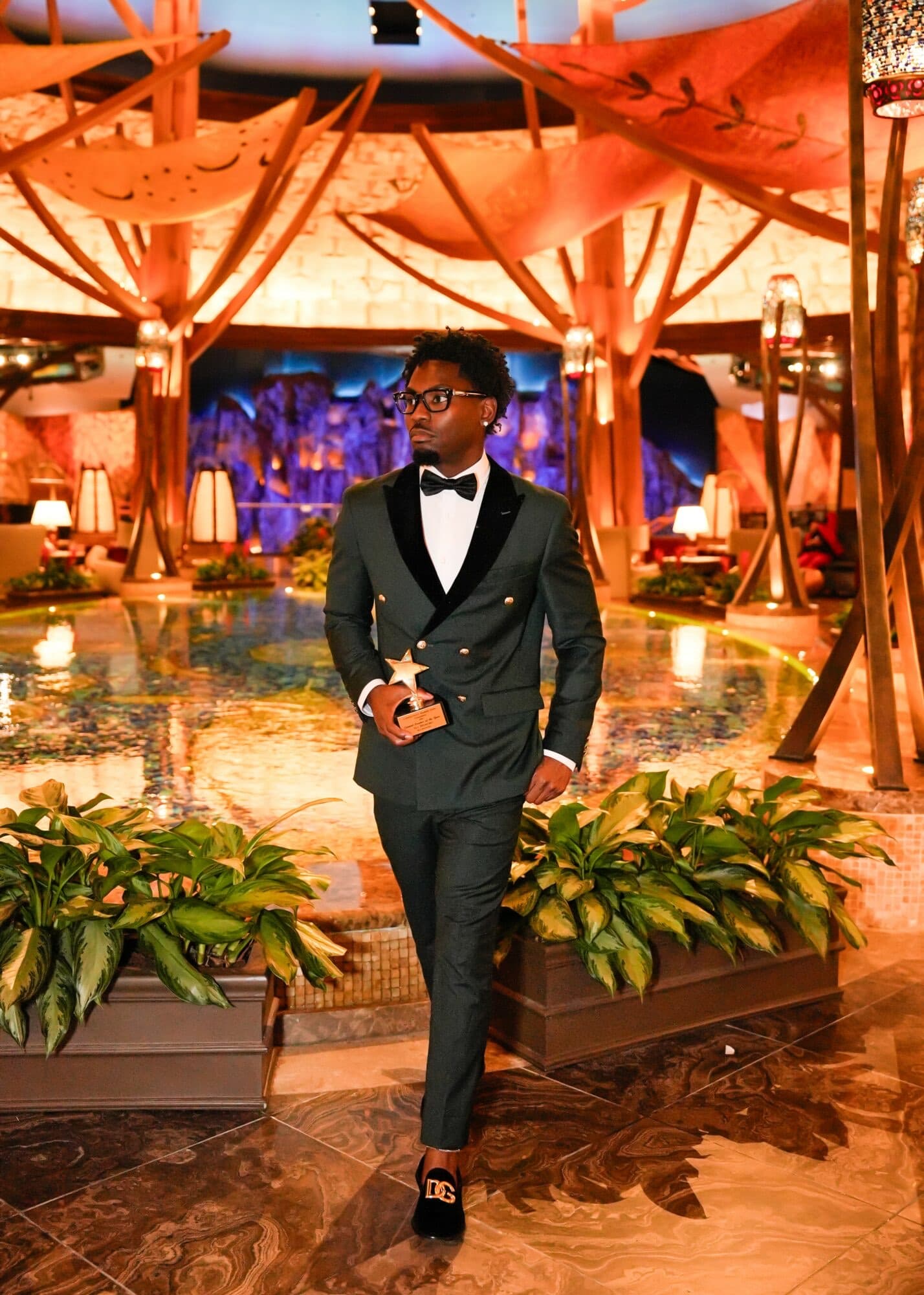Sly Huncho in a green tuxedo holding a gold star creator award in a luxury lobby