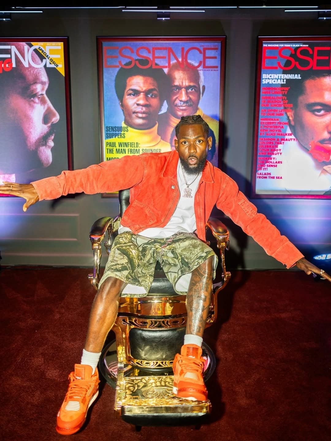 Sly Huncho seated in a gold barber chair in front of vintage Essence magazine covers