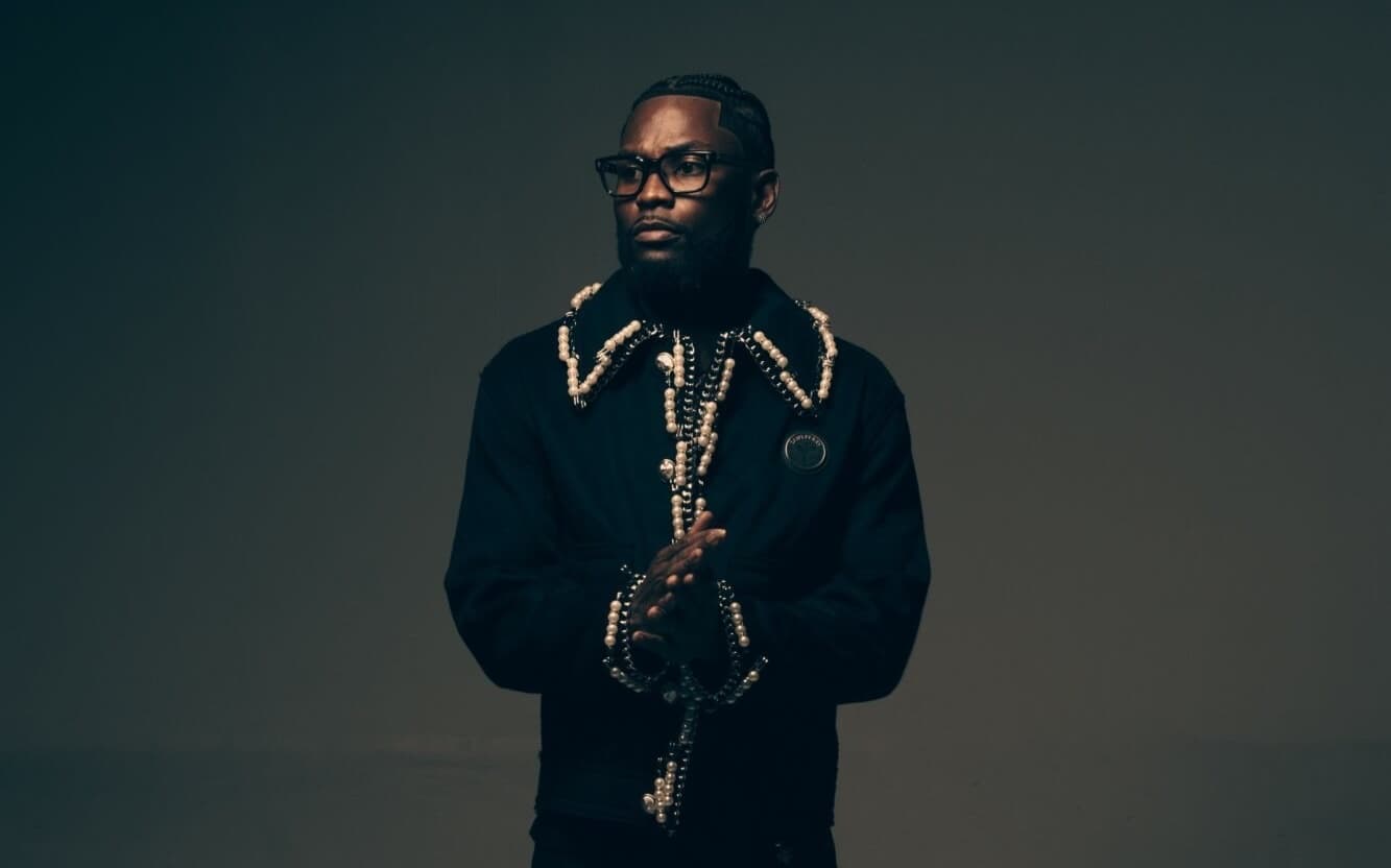Studio portrait of Sly Huncho in a black pearl-embellished jacket with hands clasped